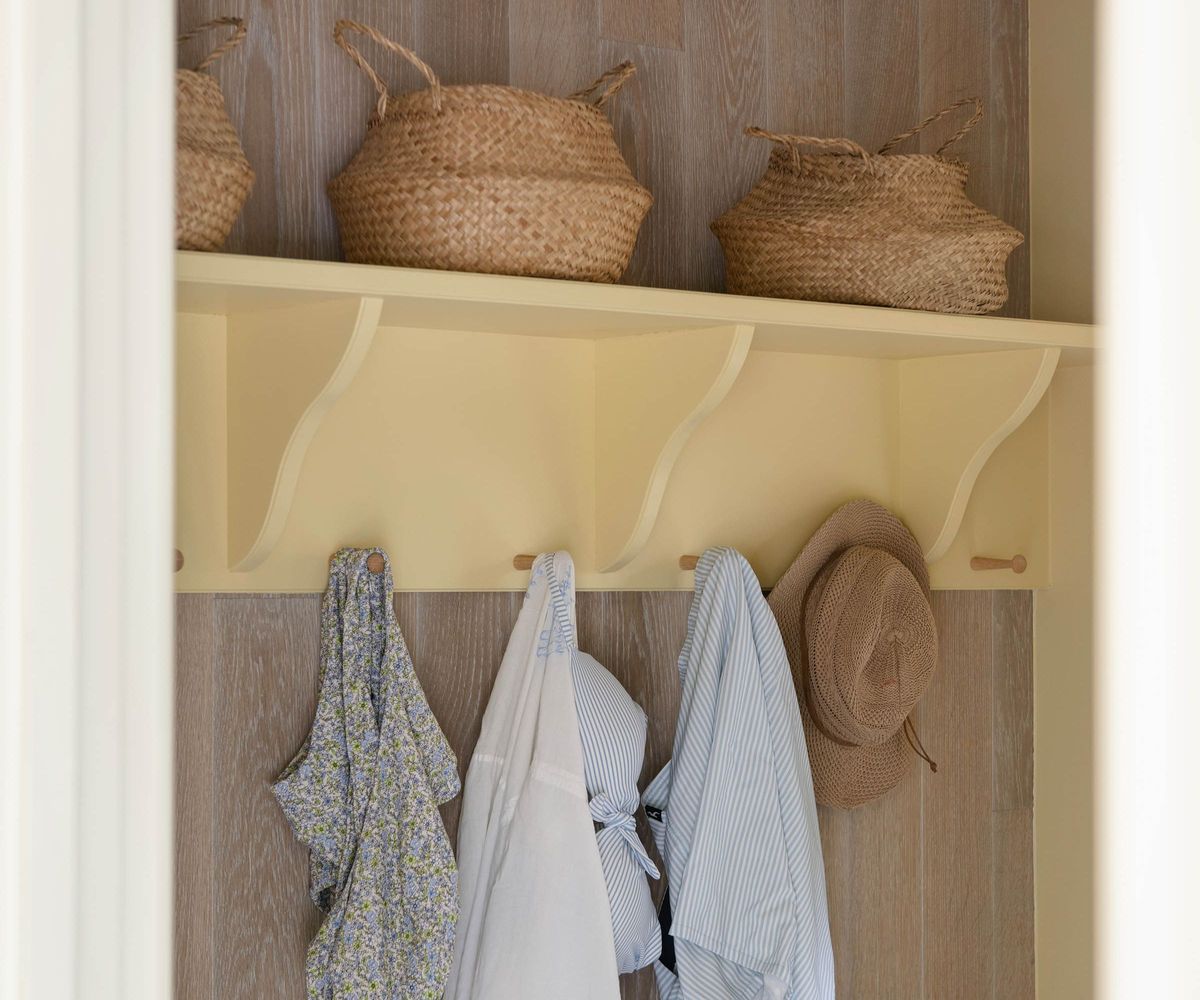 A pool house changing area with a yellow painted shelf holding seagrass baskets and wooden pegs hung with swimwear, a shirt and a straw hat.