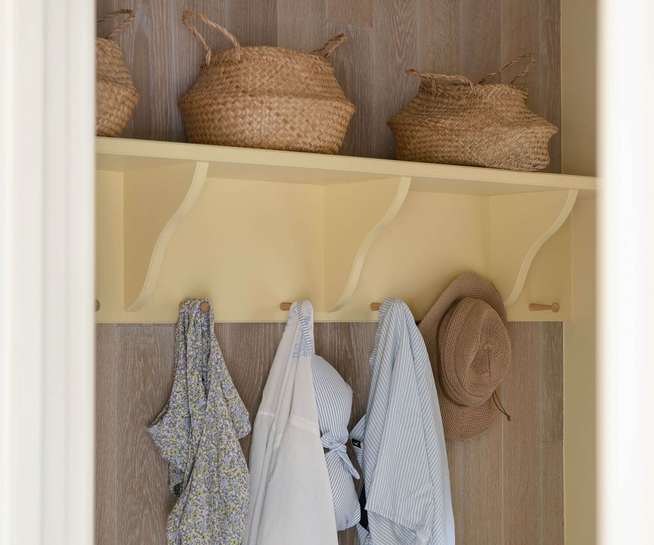 A pool house changing area with a yellow painted shelf holding seagrass baskets and wooden pegs hung with swimwear, a shirt and a straw hat.