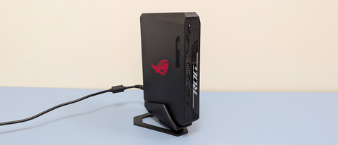 Wide angle shot of Asus ROG NUC 970 on a desk, vertical in stand, plugged in with logo glowing red