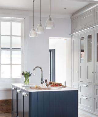 A kitchen with a dark blue kitchen island, a white surface with a silver faucet and chopping boards with bread and fruit, three glass pendant lights above it, and white cabinets to the right