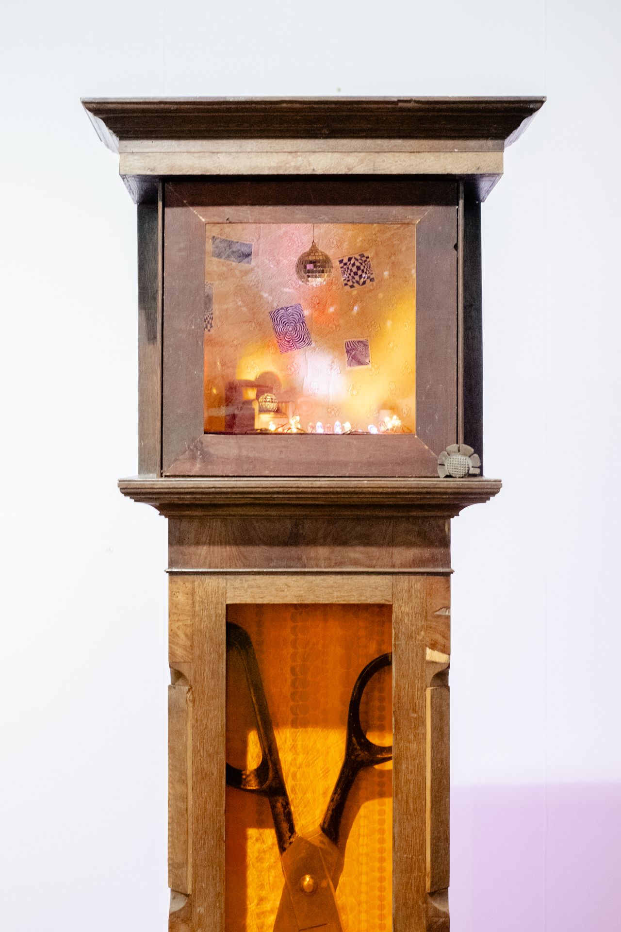 A wooden clock part of an art installation has been repurposed into a miniature display of a warmly lit living room featuring a disco ball, quirky posters, and a retro-inspired atmosphere, with the lower part of its tower filled with a giant pair of scissors.