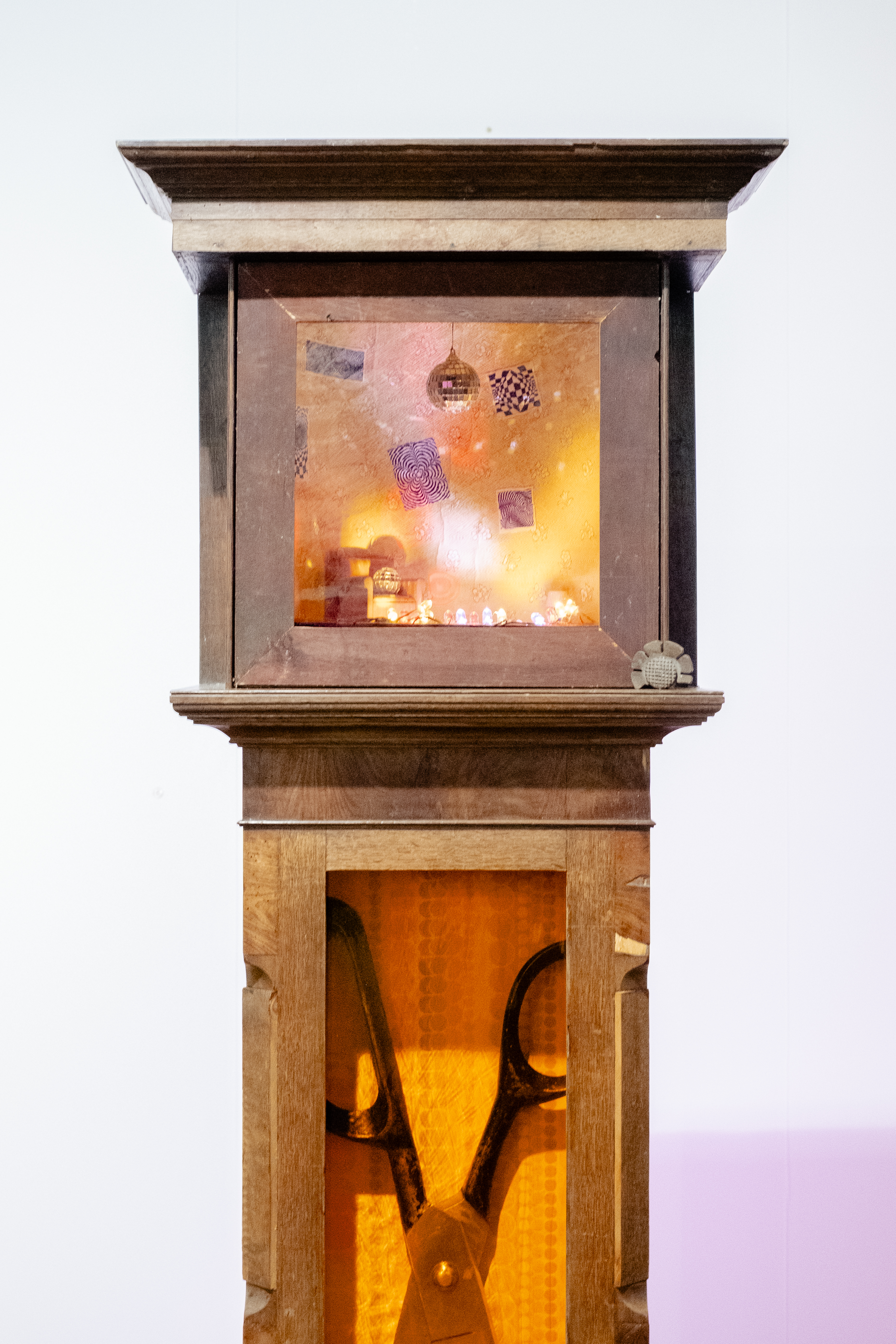 A wooden clock part of an art installation has been repurposed into a miniature display of a warmly lit living room featuring a disco ball, quirky posters, and a retro-inspired atmosphere, with the lower part of its tower filled with a giant pair of scissors.