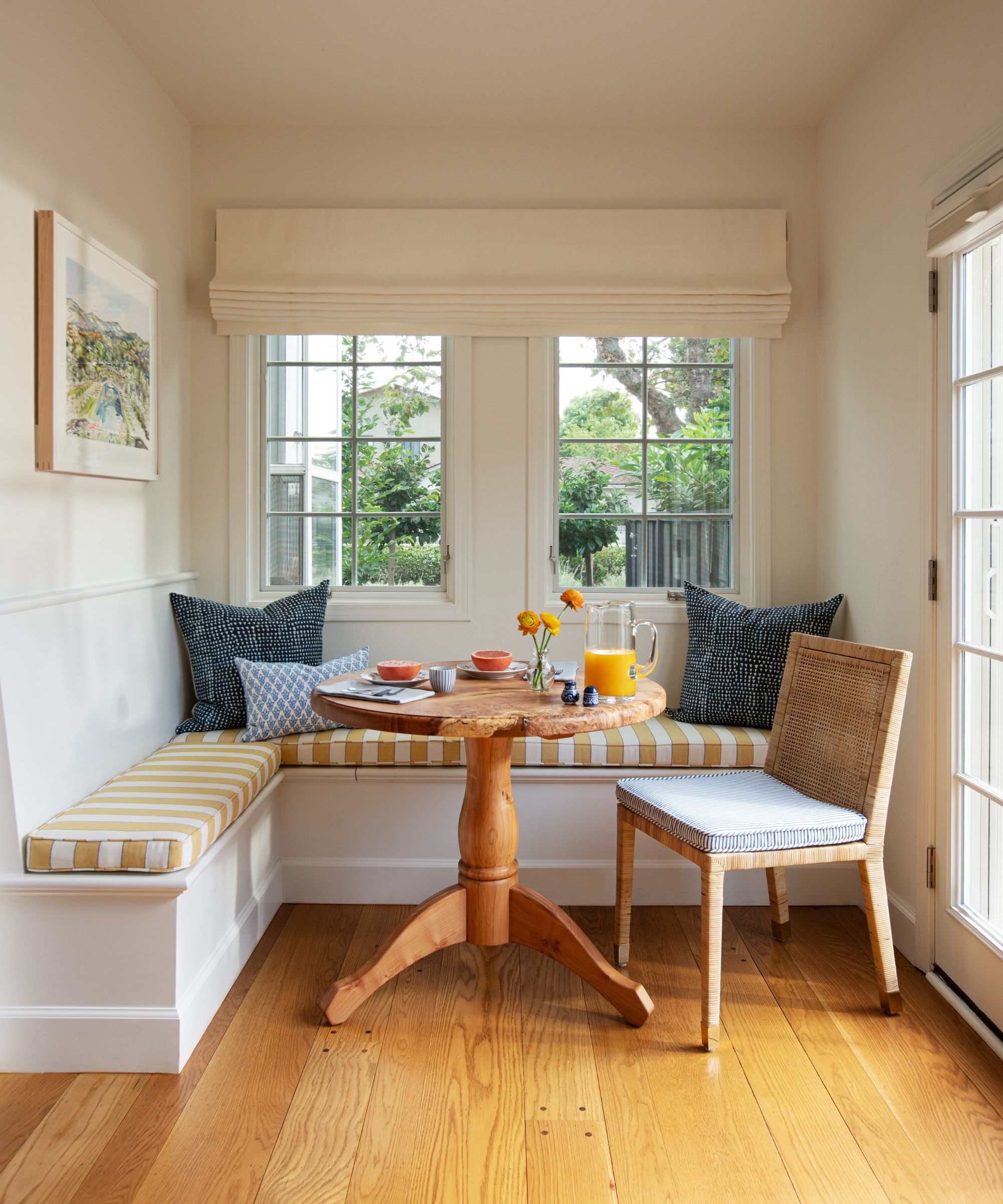 A breakfast nook in front of a window with white walls, a striped ochre corner bench, and a small, round wooden table with a single chair.