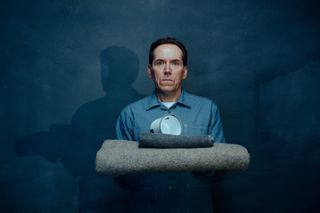 A key art shot from Professor T season 3 showing Professor Jasper Tempest (Ben Miller) in a grey prison overall standing against a charcoal-coloured backdrop. He is holding a folded prison-issue grey woollen blanket in his arms, and on top of that is a small folded black towel and a large white mug. His expression is blank, but slightly startled.