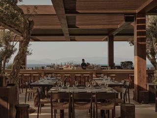 A bar tender shakes up a cocktail in a modern rustic outdoor restaurant filled with trailing plants and sweeping Mediterranean views.