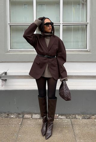 a photo of a sweater dress outfit with a woman wearing a brown mini turtleneck dress with a brown blazer, brown belt, brown tights, brown knee-high boots, and a brown handbag