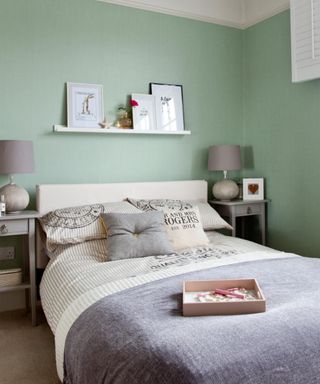 A bedroom with green walls, a white wall shelf with framed prints, a white bed with gray bedding and white and black pillows, a rectangular wooden tray on top of it, and two gray nightstands with round lamps each side