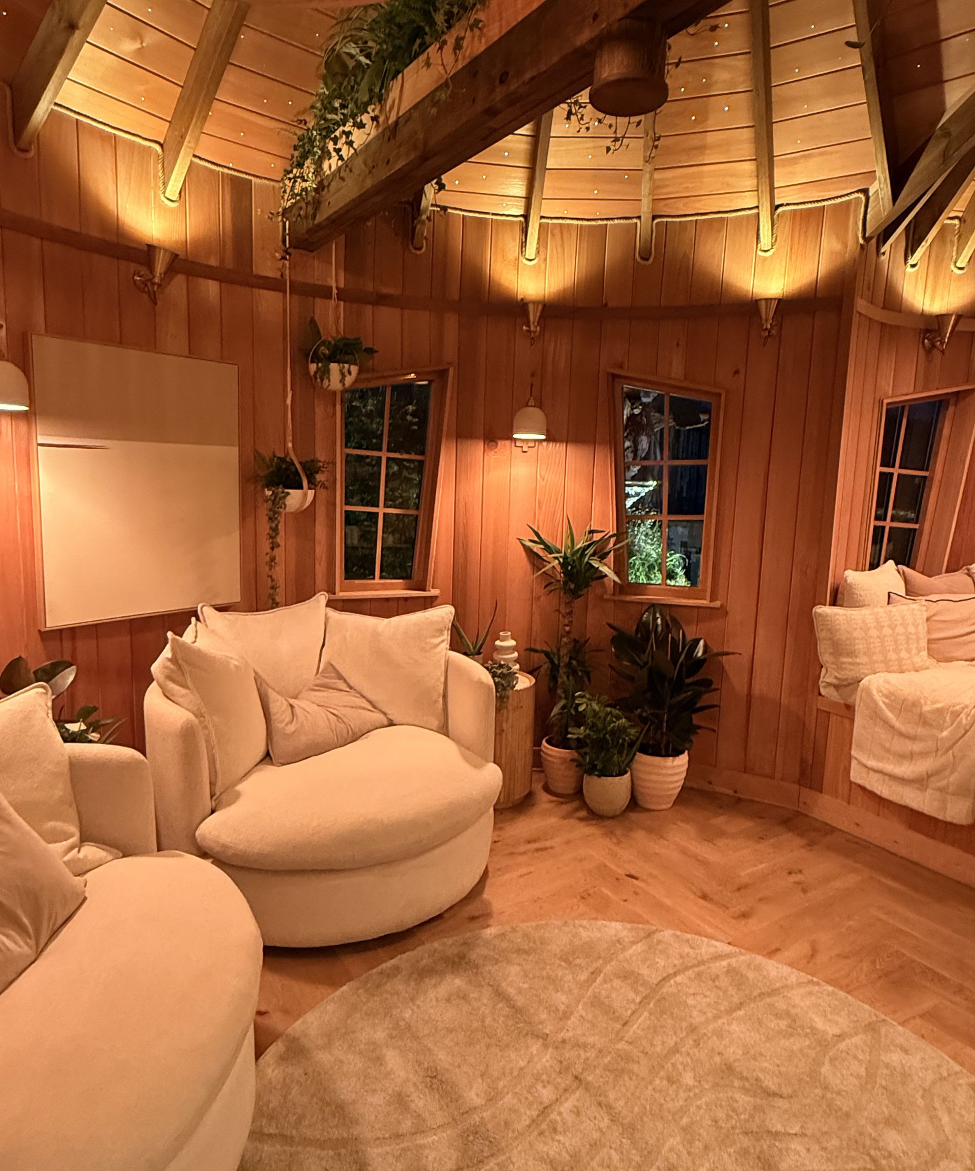 Cosy treehouse interior with warm wood-panelled walls, plush boucle armchairs, and layered neutral textures. Ambient lighting and potted greenery create a calming retreat, complete with a built-in daybed and curved ceiling beams strung with fairy lights.