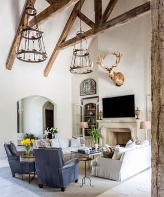 Large traditional living room with vaulted ceiling and beams, seating area positioned around a fireplace, tv mounted on wall, two low hanging chandliers