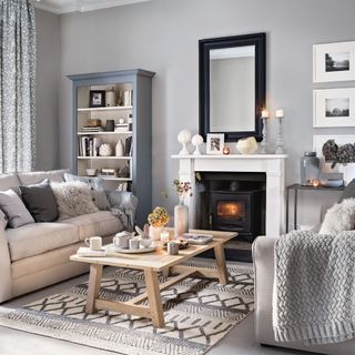 grey living room with a black mirror above a white fire place with wall-mounted picture frames and candle-accessories with a grey and cream-colored sofa with neutral and furry cushions and a wooden coffee table