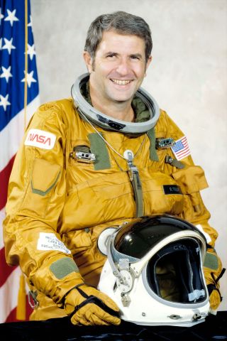 a smiling man in an orange-brown flight suit stands in front of an american flag
