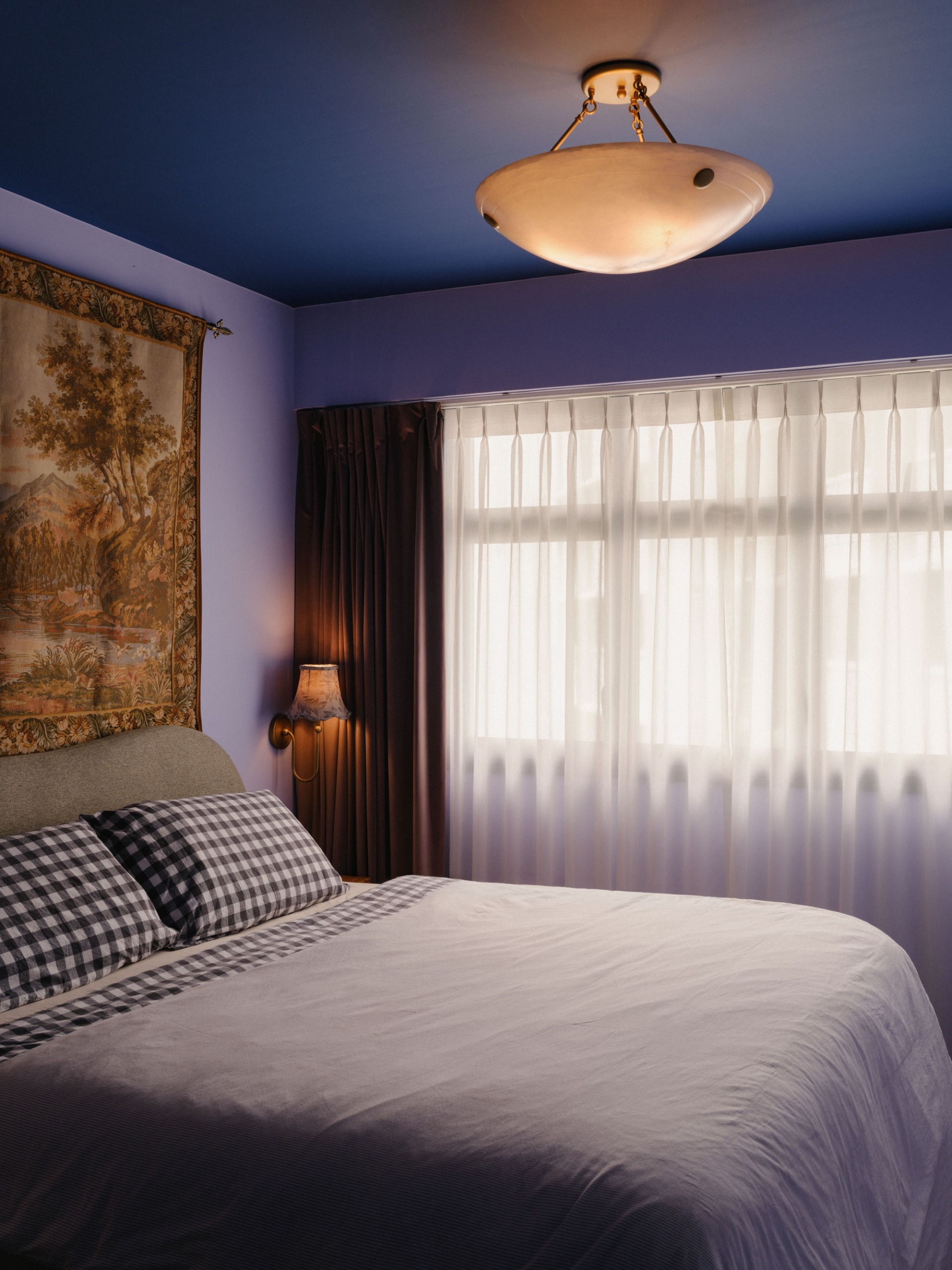 a blue bedroom with a tapestry wall hanging, checkered gingham sheets and a wall lamp
