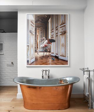 White bathroom with a copper coloured bateau bath in a wood floored room with a large painting above, wooden flooring.