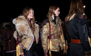 Three female models wearing looks from the Dsquared2 collection. One model is wearing a multicoloured top, black trousers with black detail and a brown sleeveless fur jacket. Next to her is a model wearing a brown and green camouflage style jacket with a brown belt. And the third model is wearing a dark coloured belted piece featuring lace, tassels and metallic embellishments. Two of the models are wearing pieces that feature yellow and red tassels