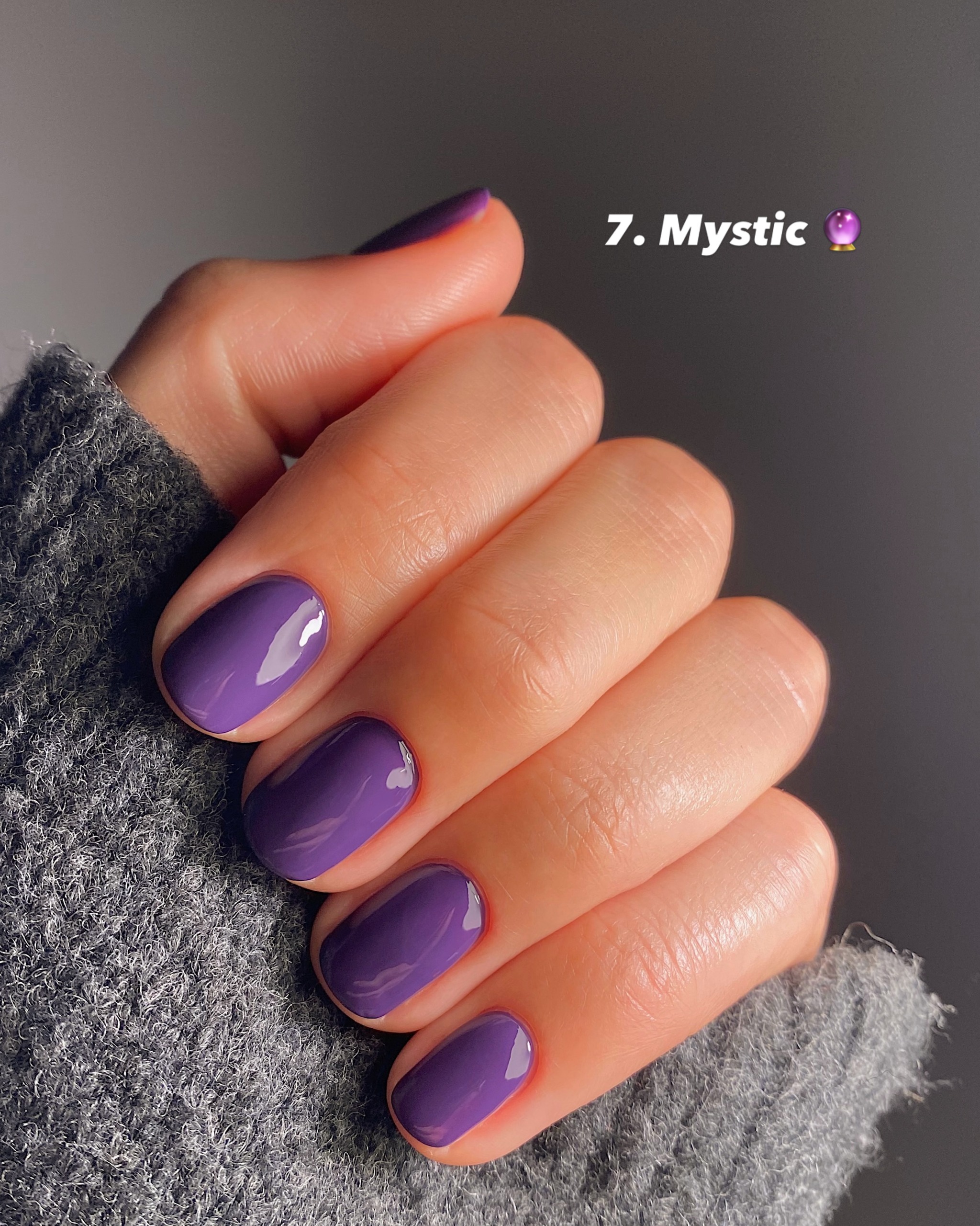 Short glossy purple nails, hand tucked into a gray sweater sleeve