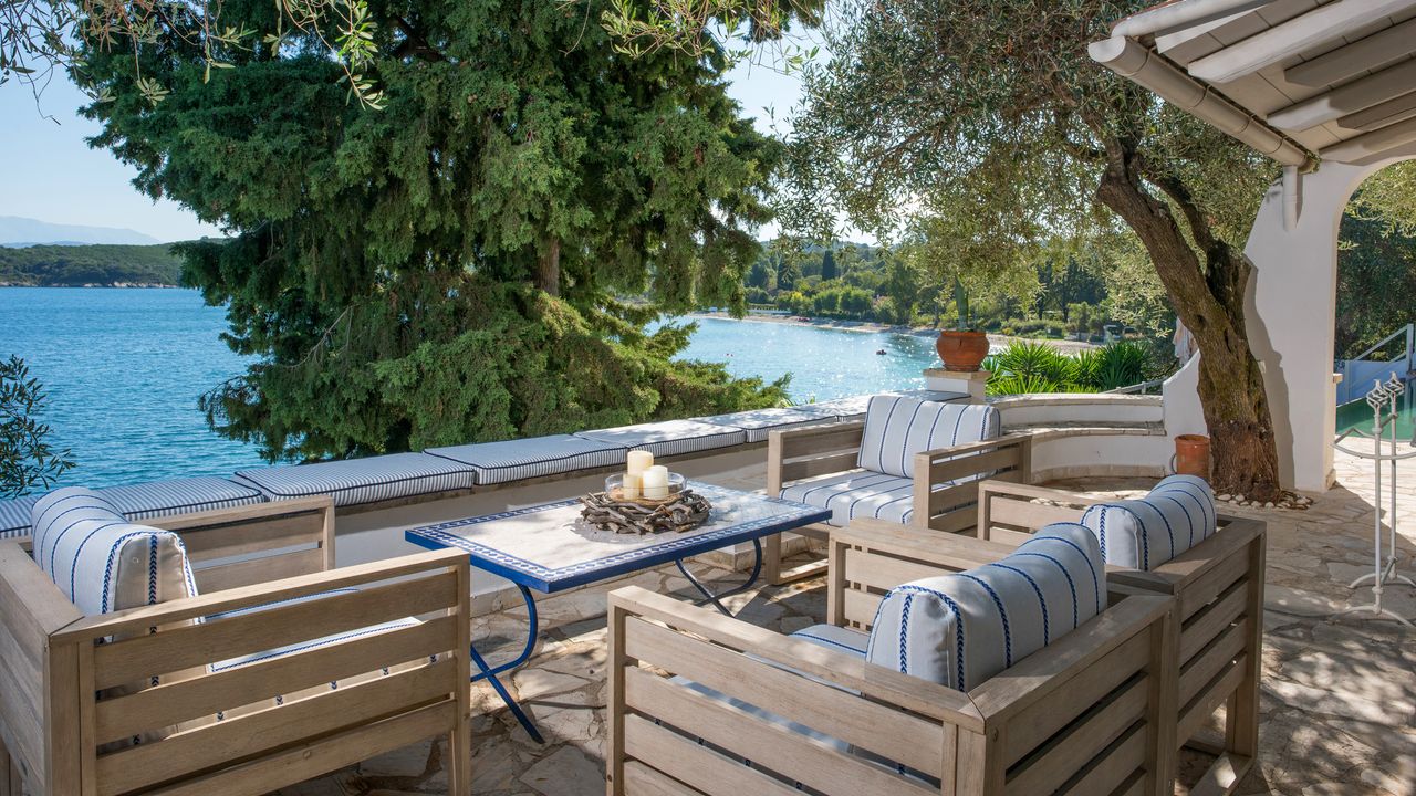 A seating area at a Corfu villa with blue and white-striped cushions on wooden armchairs, and a sea view