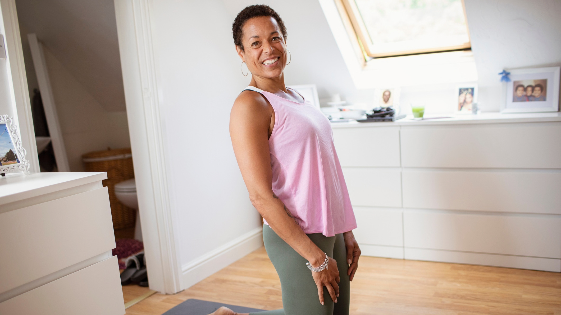 Woman in sportswear kneels on the floor in a domestic setting, she is smiling at the viewer 