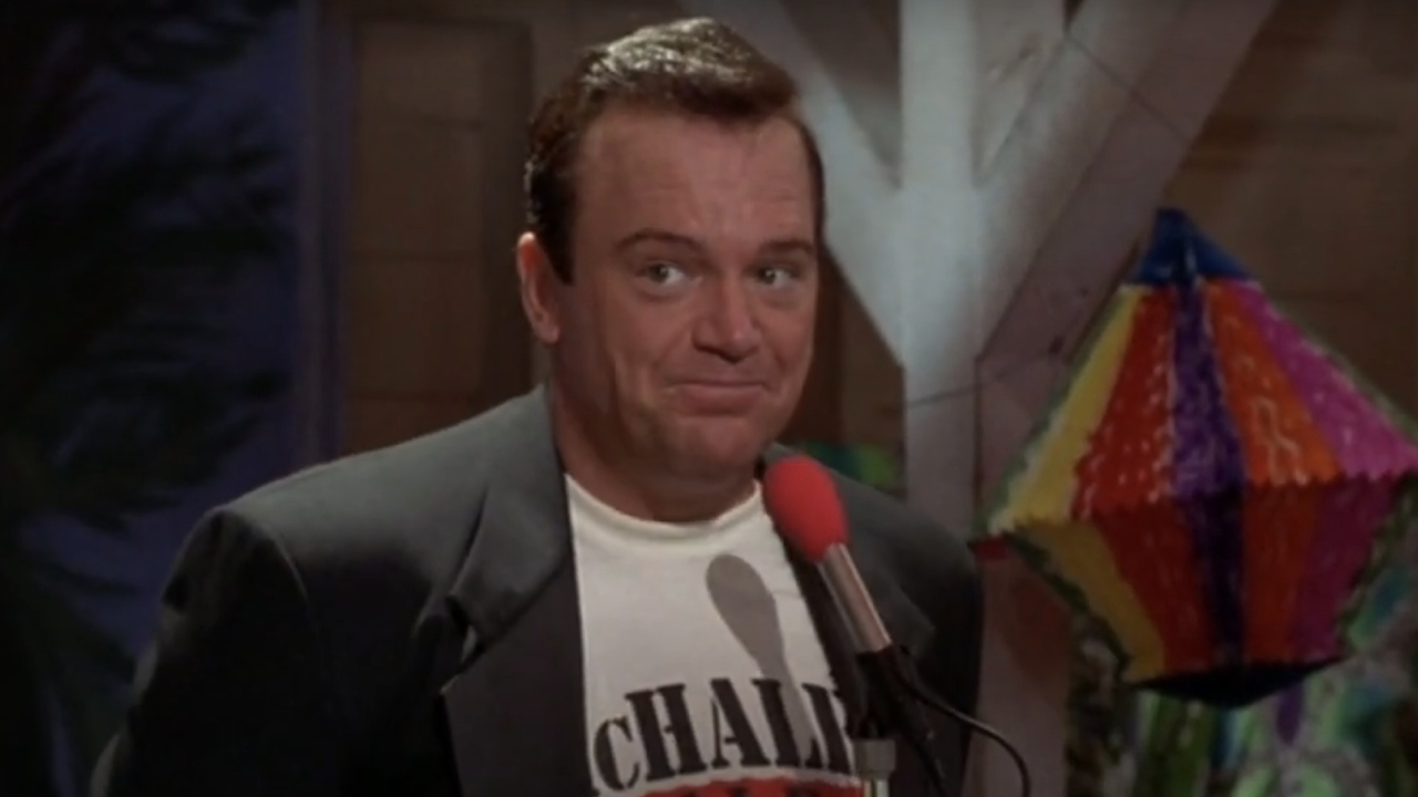 Tom Arnold smirking in front of a microphone in McHale's Navy