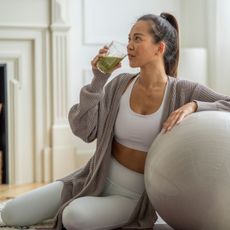 A woman drinking a smoothie in workout gear leaning on a workout ball