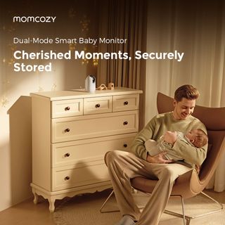 A parent holds their baby in a rocking chair seated in a beige-toned nursery with a Momcozy baby monitor camera sitting nearby on a dresser.