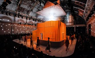 Models walking around a wallpaper house in the middle of a runway with a glitter ball illuminating the fashion show space