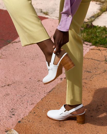 A woman wearing green pants standing on one leg adjusting her white high heel.