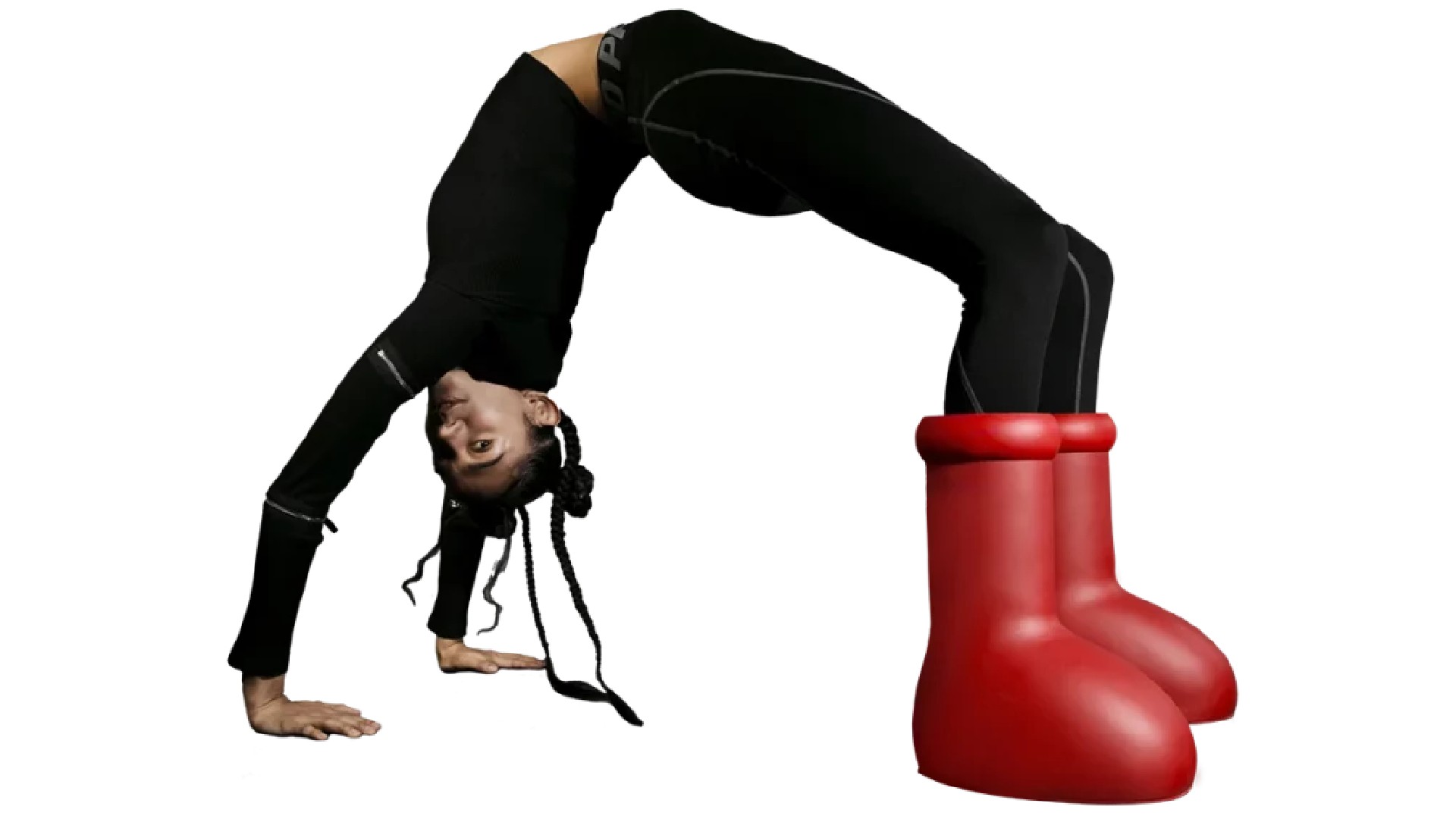 A woman in black athletic wear performs a backbridge pose while wearing oversized, bright red, cartoonish boots against a white background.