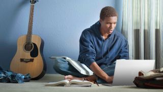 Man using Dell XPS 13 with a book open next to him, and an acoustic guitar behind him.