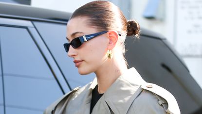 Hailey Bieber walks to a car wearing a trench coat and a pair of sunglasses plus croissant hoops