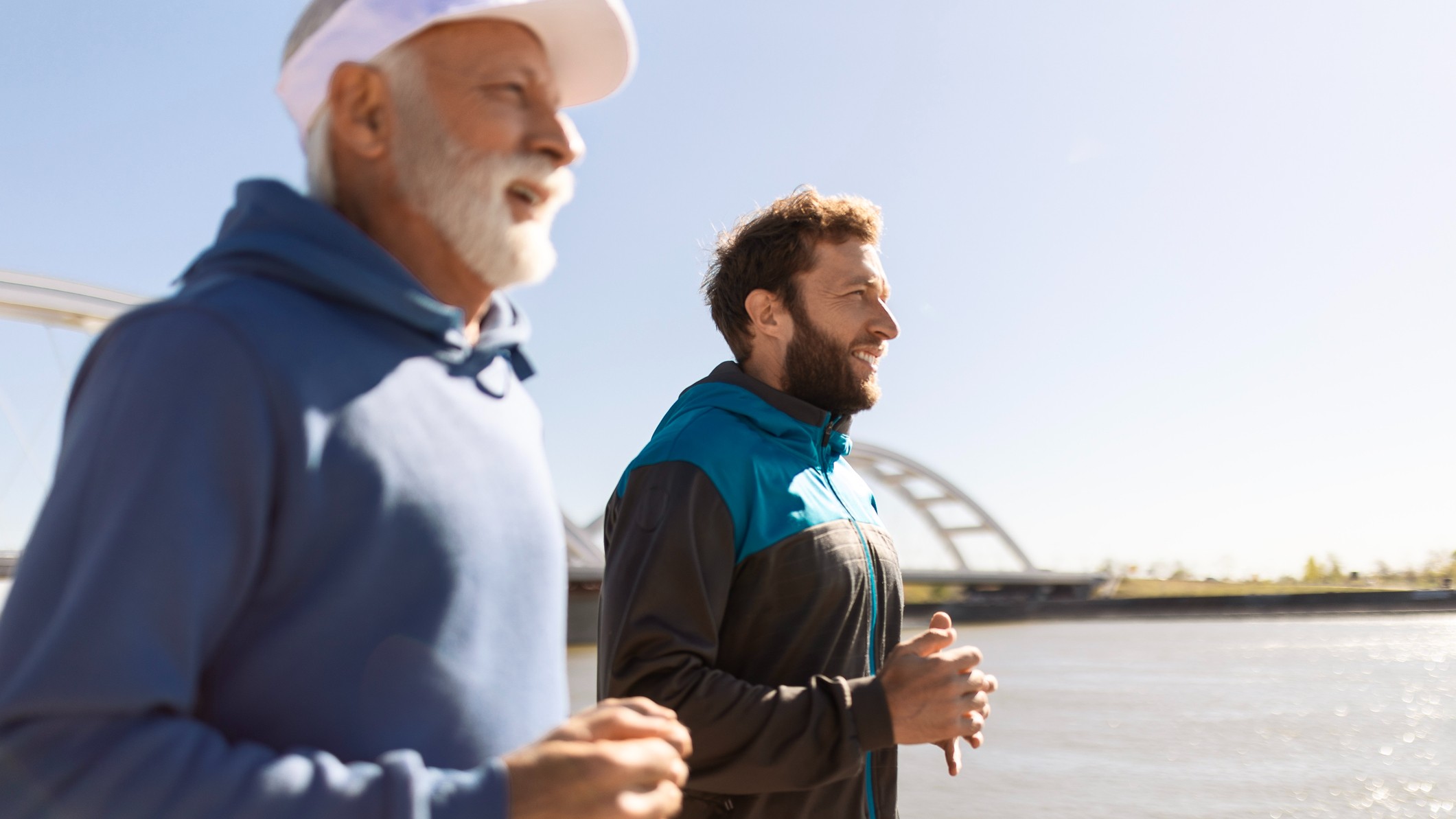 A father and son jog alongside a river. The father is an older man and the son is middle-aged.