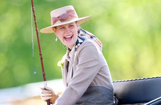 Lady Louise wearing a tan jacket and straw hat holding a horse whip and laughing in a carriage