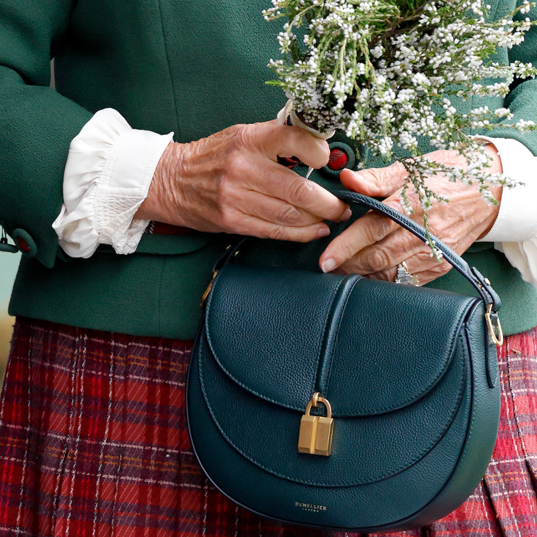 A close up of Queen Camilla's red tartan skirt and hands holding a green Demeillier bag
