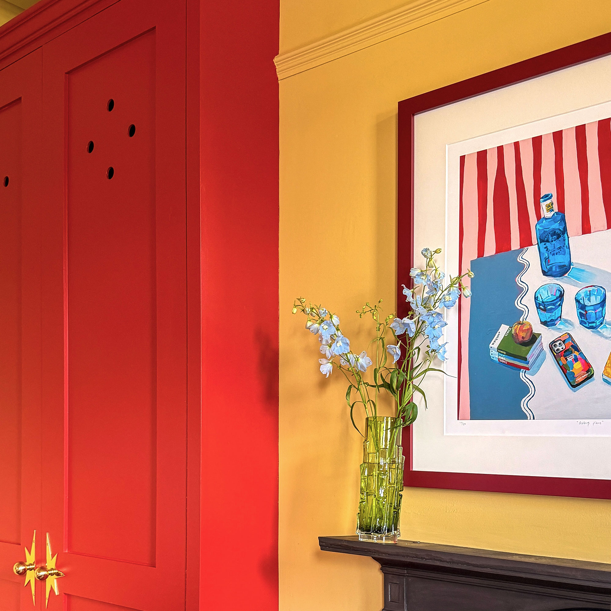 Yellow room with red wardrobe, colourful painting above mantlepiece with glass vase of blue flowers