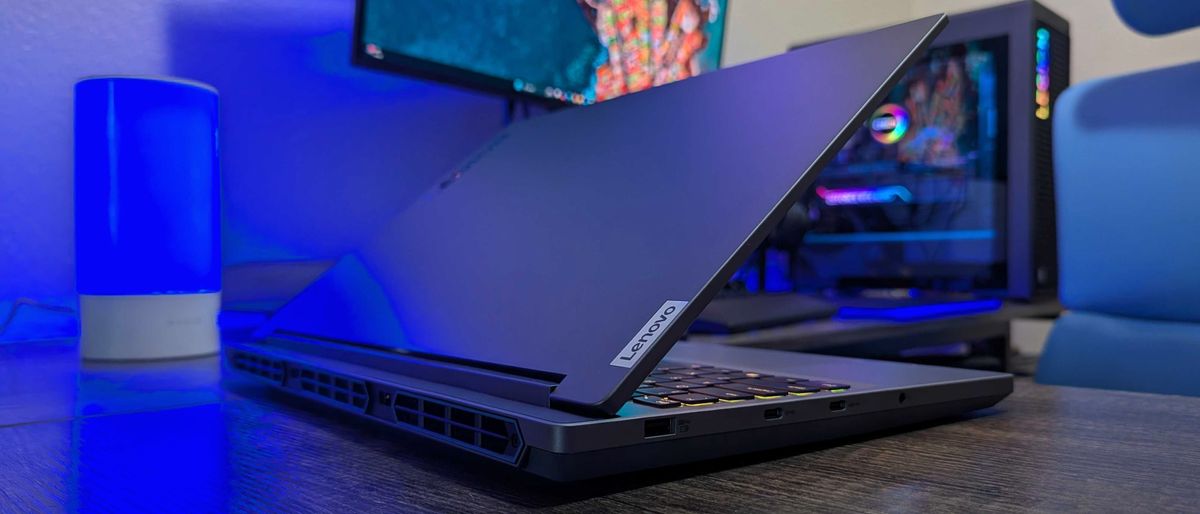 A Lenovo Legion 5i (Gen 9) gaming laptop on a desk with a blue backlight, facing away at an angle with its lid open to show its rear and right side ports, a bit of its RGB-lit keyboard, and the &quot;Lenovo&quot; logo on the lid.