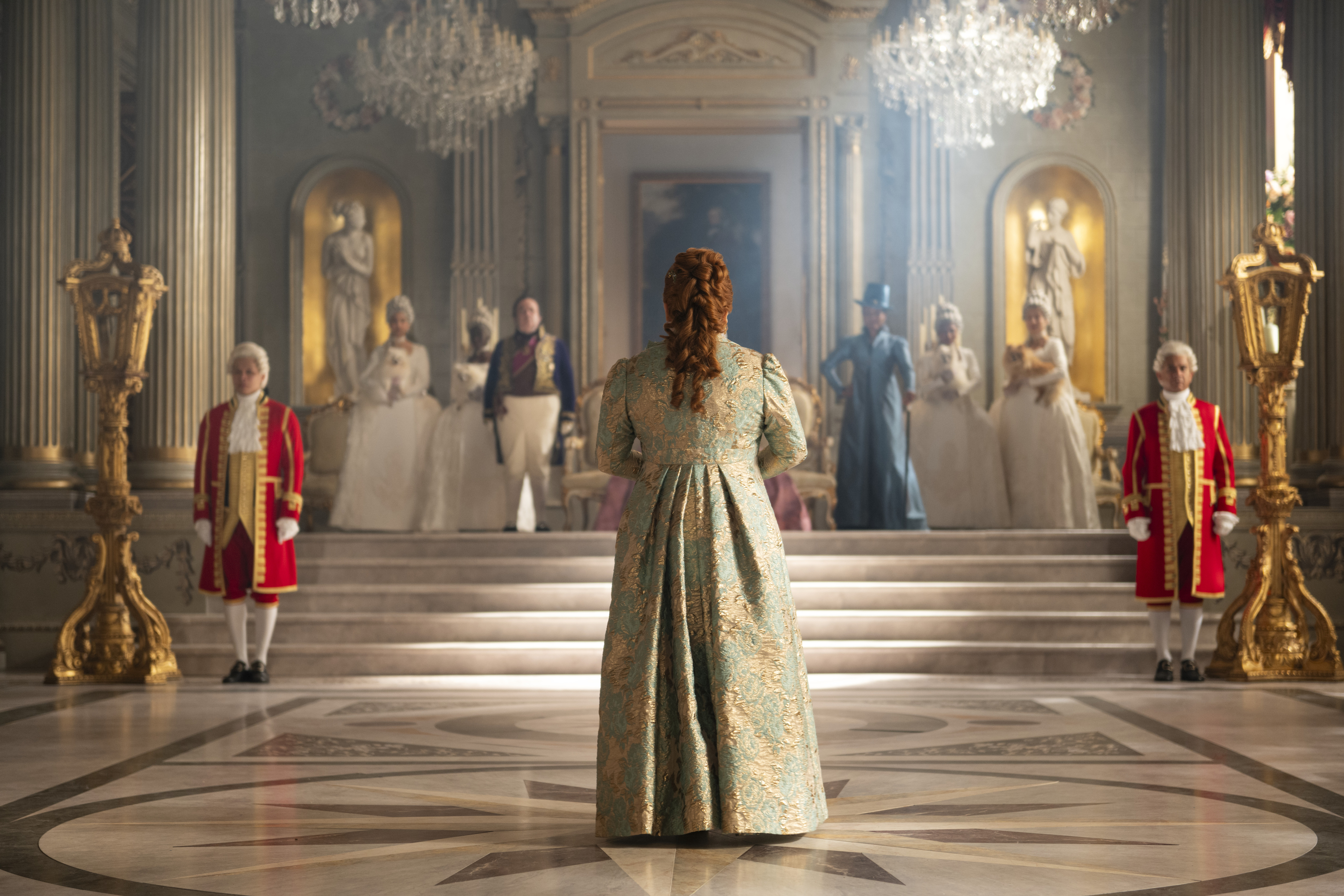 Hugh Sachs as Brimsley, Nicola Coughlan as Penelope Bridgerton, Adjoa Andoh as Lady Danbury standing in the queen's court before her throne as penelope approaches the queen