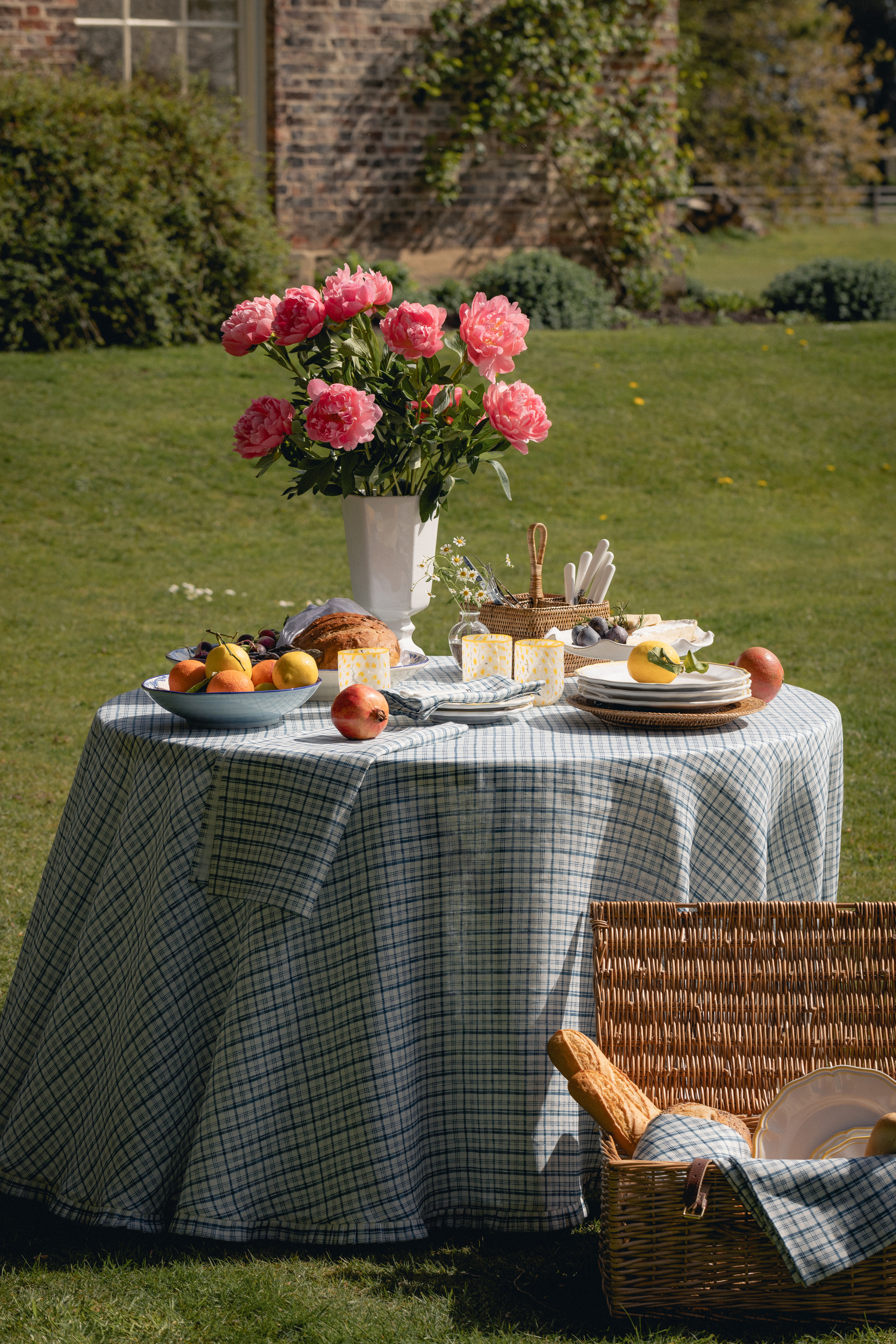 an outdoor round table with a checkered tablecloth, set with pink flowers, citrus, yellow glasses, and a cutlery basket