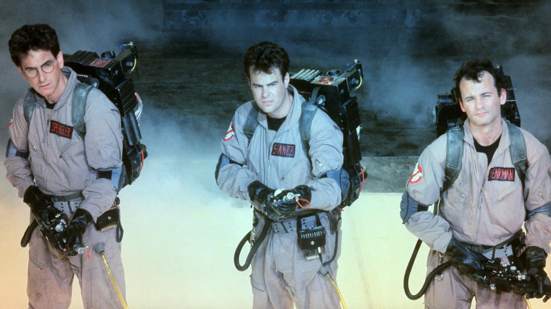 Screenshot from the movie Ghostbusters. A trio of Ghostbusters. Three men all wearing beige jumpsuits with giant black backpacks with a gun/nozzle attached to it. They are standing in a fog and aiming their weapons.