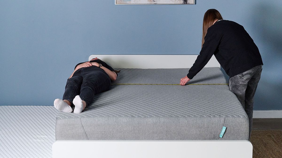 This images shows our sleep writer Ruth Jones testing the edge support of the Siena Memory Foam Mattress. The Siena Mattress is on a bed frame in our sleep studio. To the left, Ruth Jones lies on the very edge of the mattress where she seems well balanced (a crash pad is placed on the floor to the left of the mattress.) To the right, sleep writer Frances Daniels measures the distance between Ruth's hips and the furthest edge of the mattress.