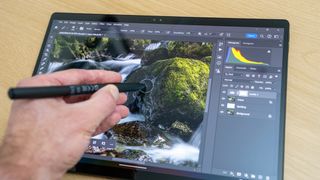 A hand using a stylus pen on the screen of an Asus ProArt PZ13 laptop on a wooden table