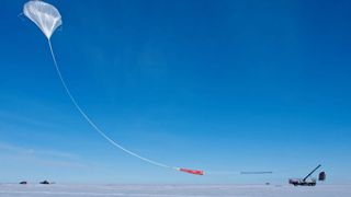 a large white scientific balloon launches into a blue sky from a flat, snow-covered patch of ground.