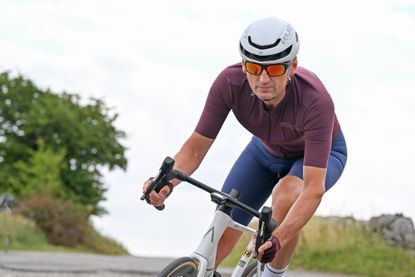 Man wearing a purple jersey, blue cycling shorts and a white helmet riding a white bike downhill head on