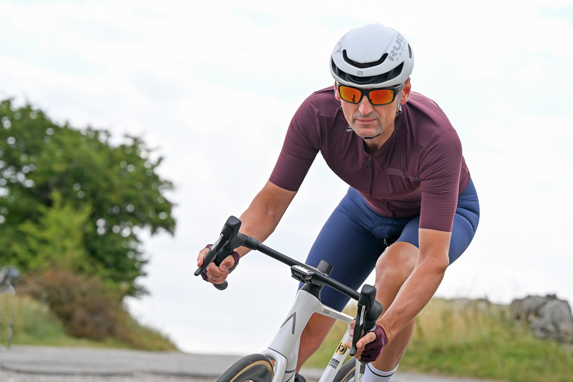 Man wearing a purple jersey, blue cycling shorts and a white helmet riding a white bike downhill head on