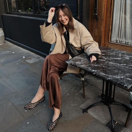 British style influencer Rachael Keegan sits at an outdoor marble cafe table wearing a cropped trench coat, brown satin pants, and studded black mesh ballet flats