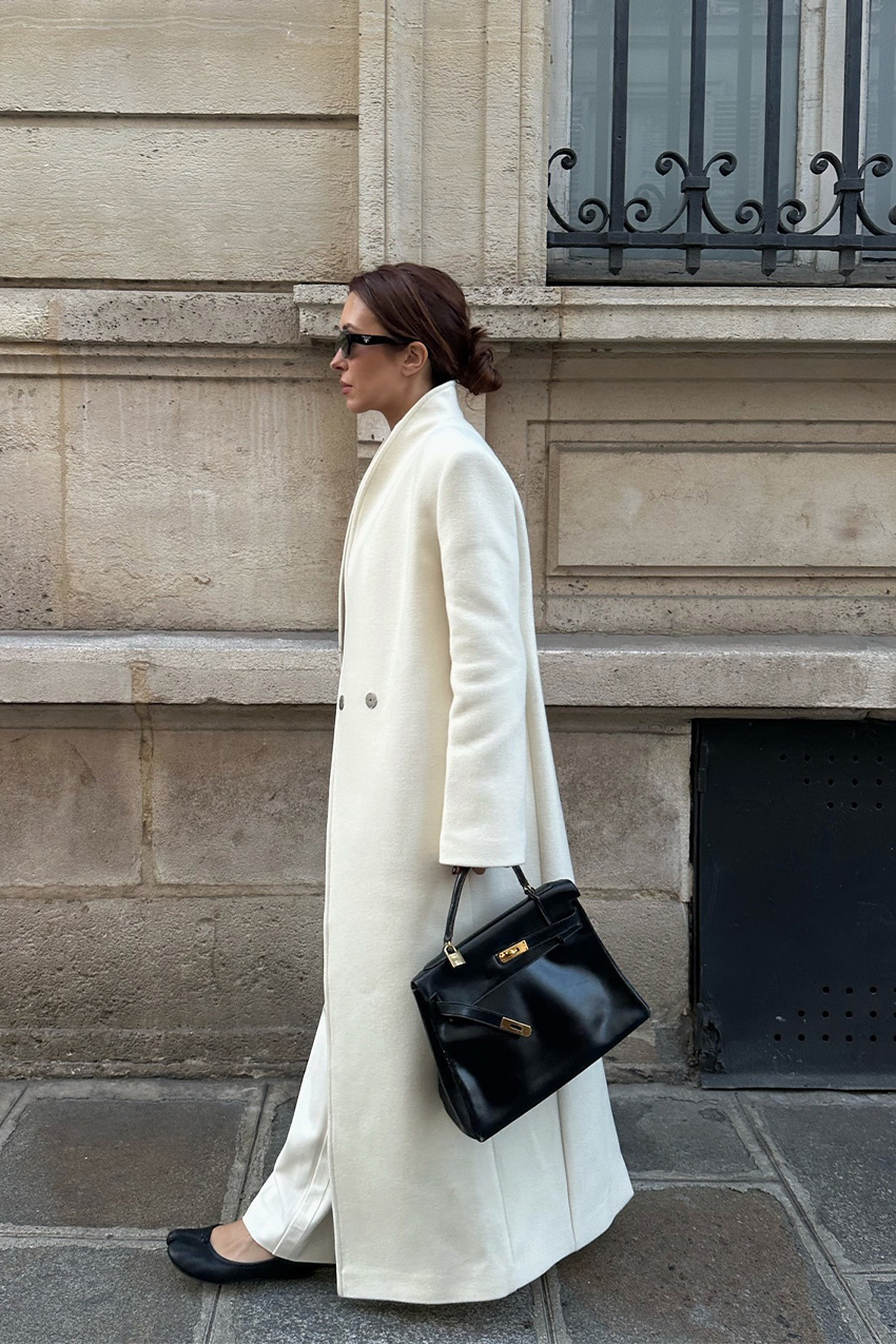 A woman walking down the sidewalk wearing black sunglasses, a long white maxi coat, white trousers, black Maison Margiela Tabi ballet flats, and a black Hermes handbag.