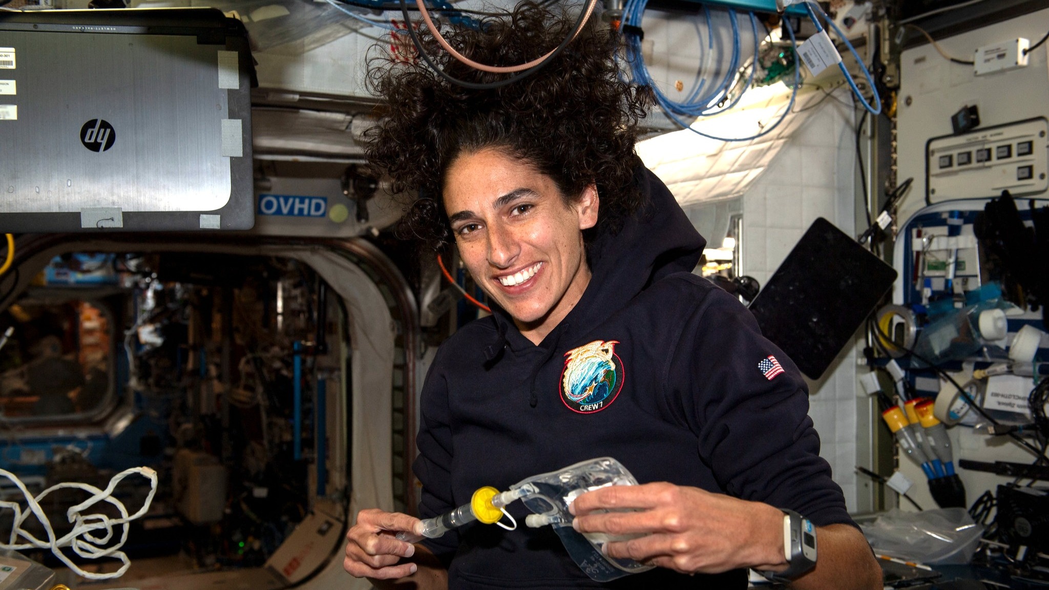 jasmin moghbeli holding a small water bag in a space station module