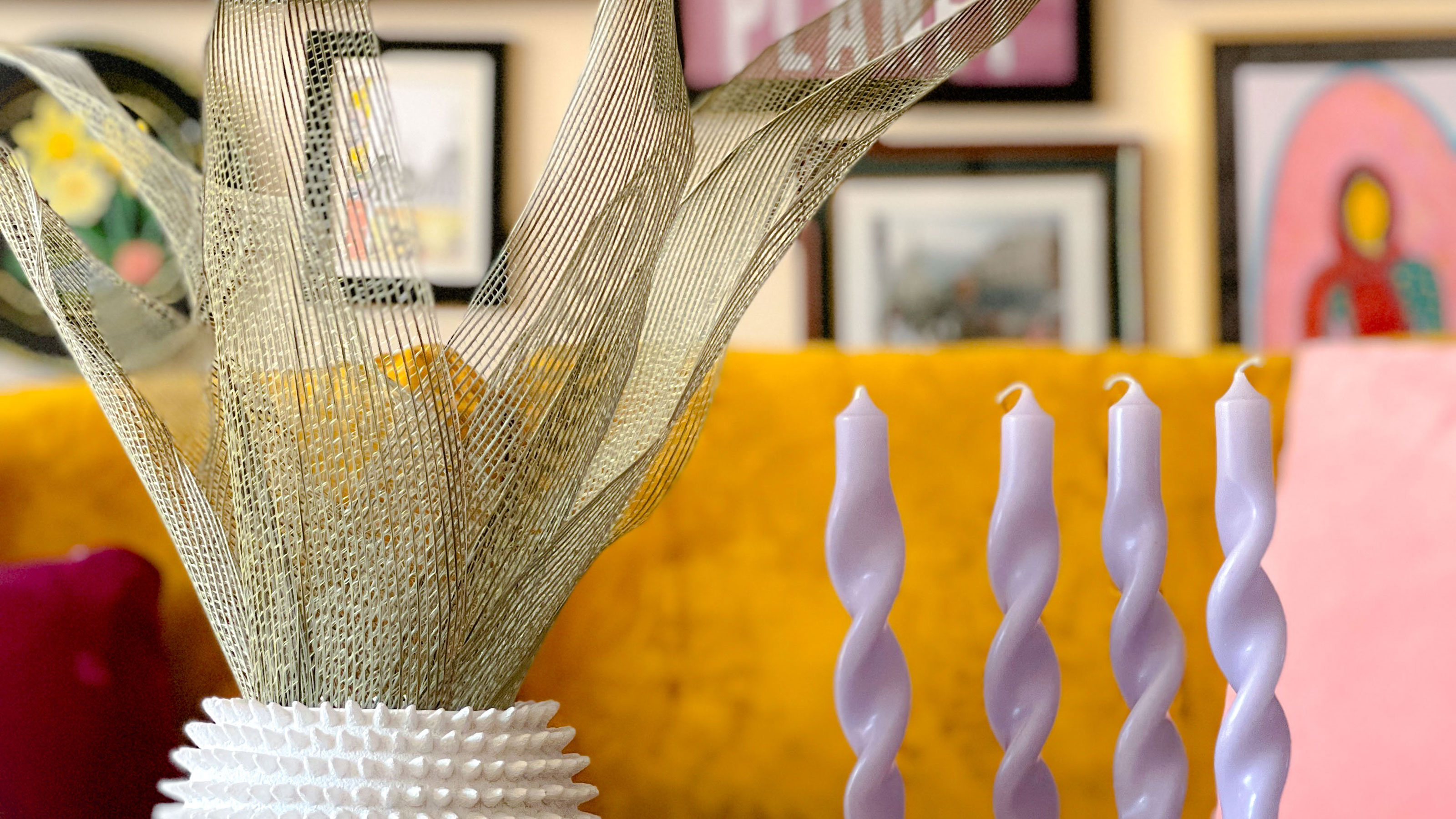 Vase with faux leafy plant beside four lilac candles, in front of mustard sofa and colourful gallery wall
