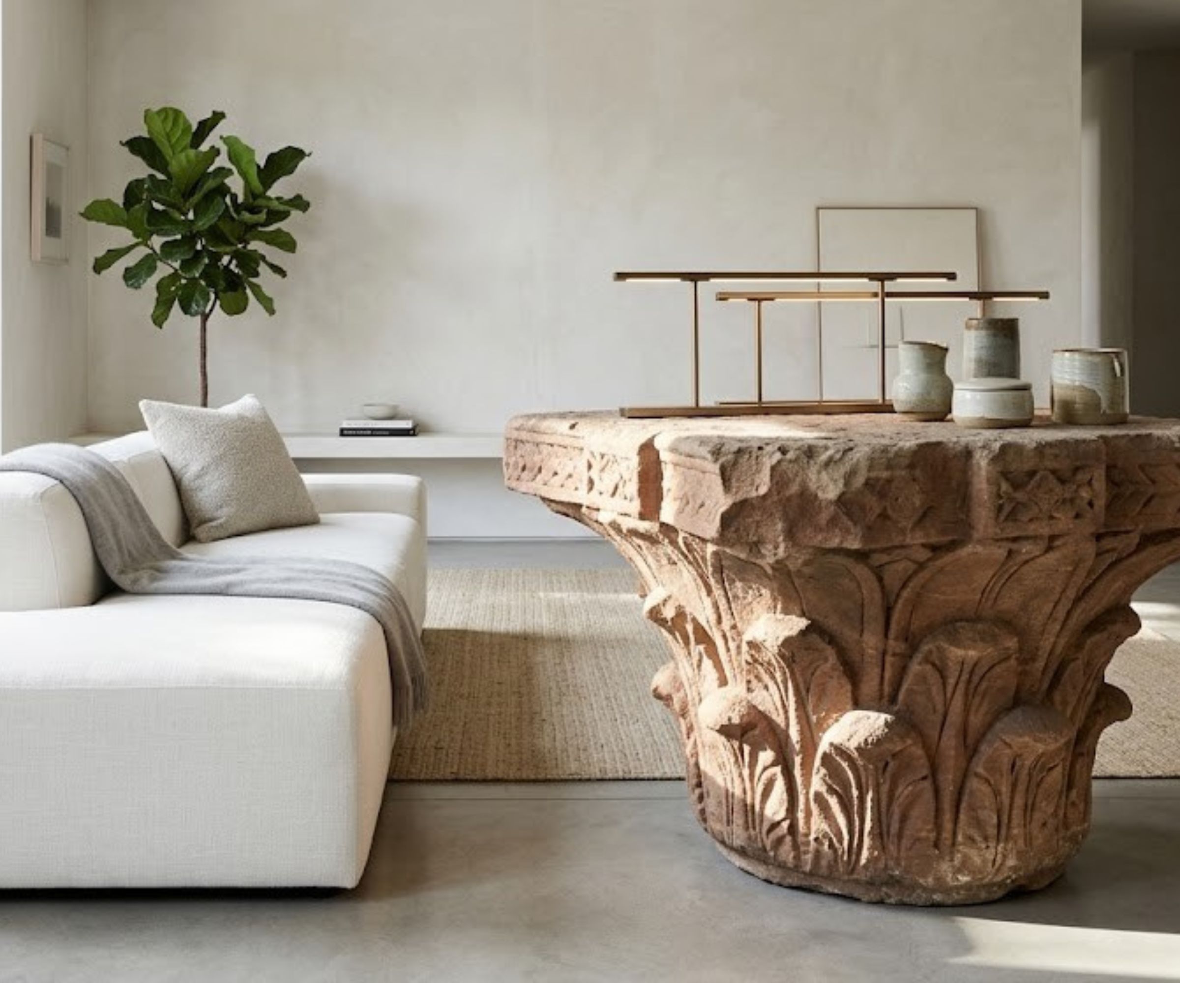 Minimalist living room with limewash walls, concrete floor, a jute rug on top, a white sofa, and a large antique Indian column made from stone used as a coffee table.