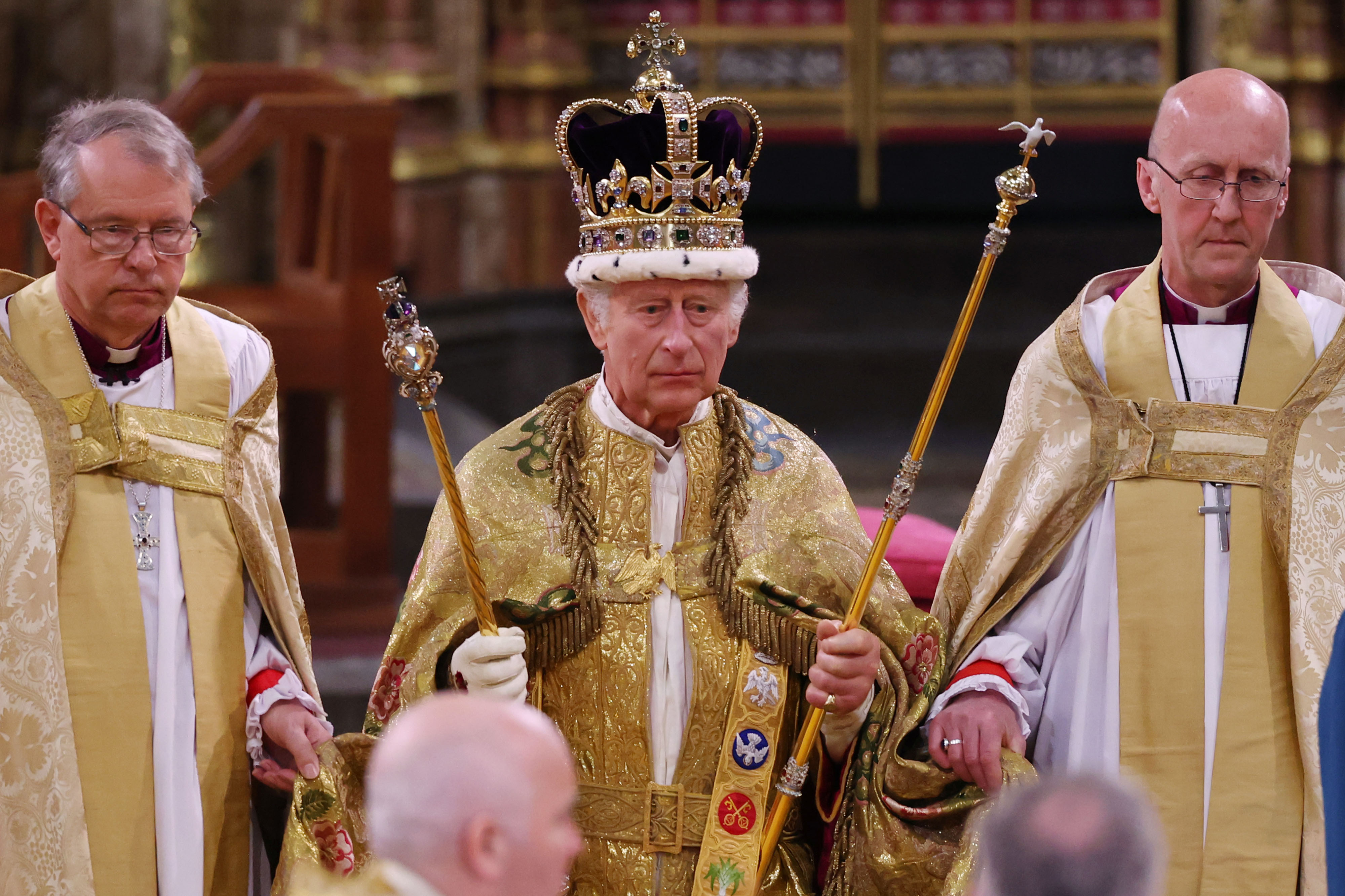 King Charles at his coronation in Westminster Abbey wearing a crown and regal attire, holding sceptres, standing between two men in gold robes.