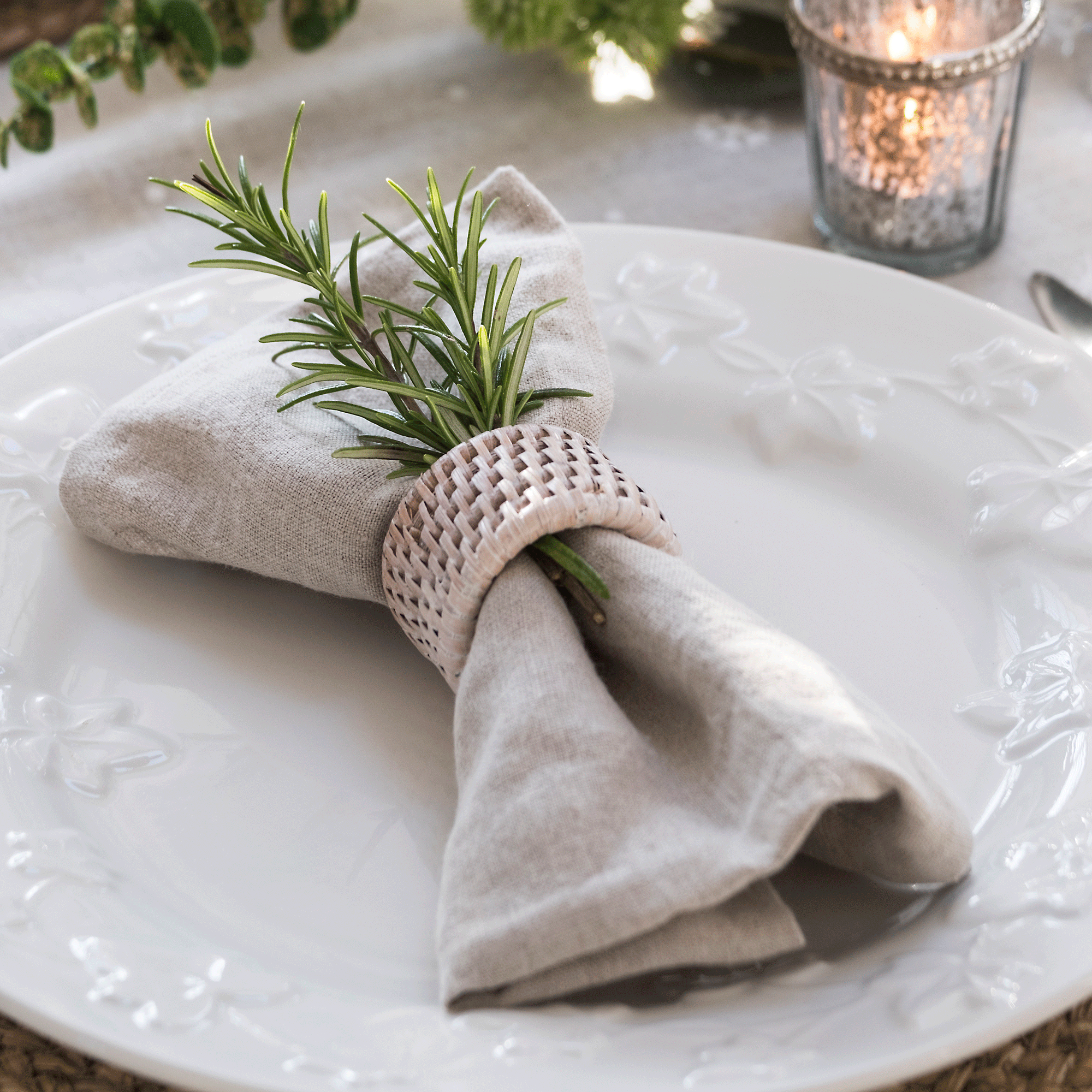 a festive neutral place setting with a linen napkin secured with a wicker napkin ring and a spring of rosemary