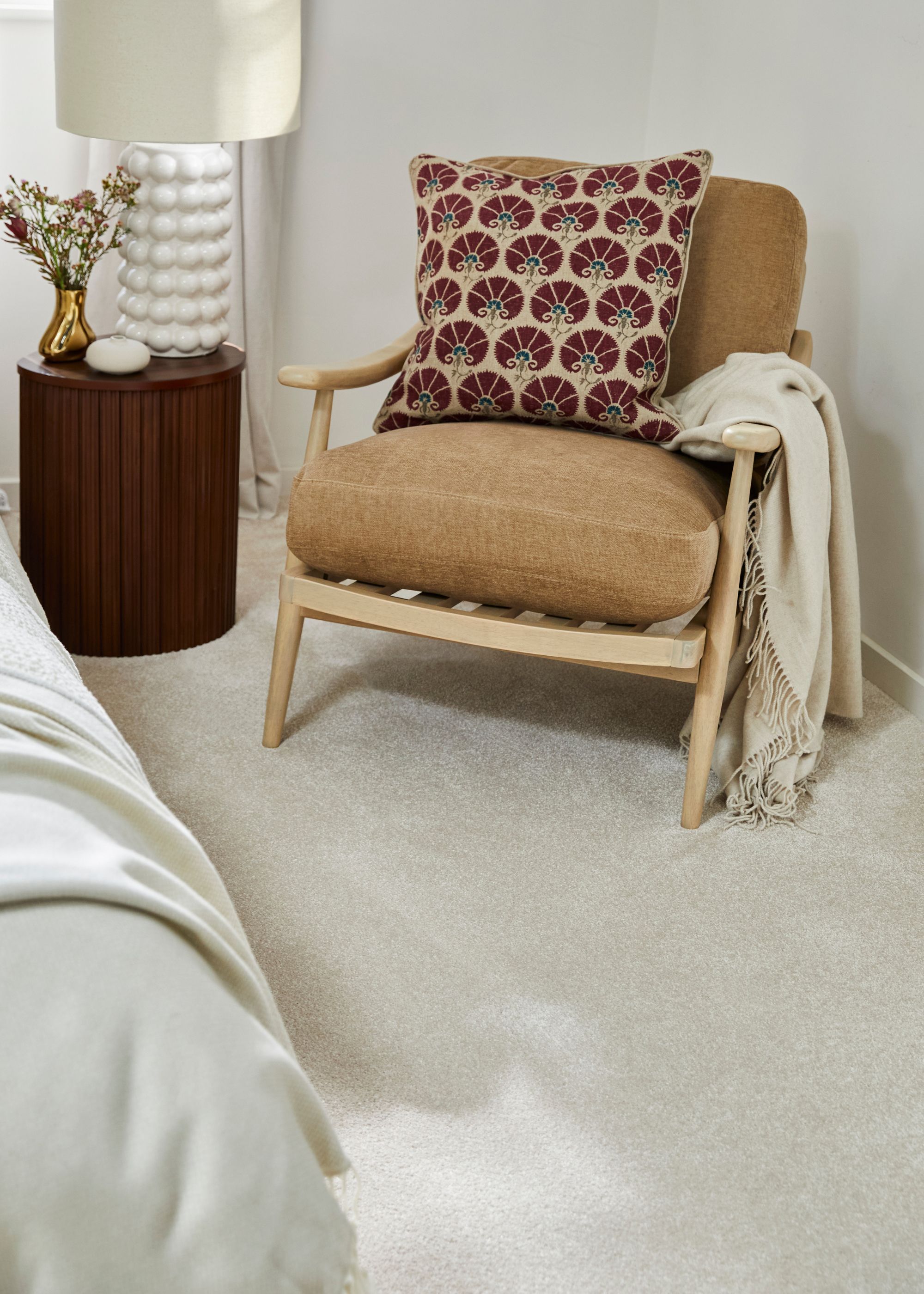 A neutral colored carpet with a chair. The chair has a cushion on top of it. There is also a side table with a lamp and a vase.